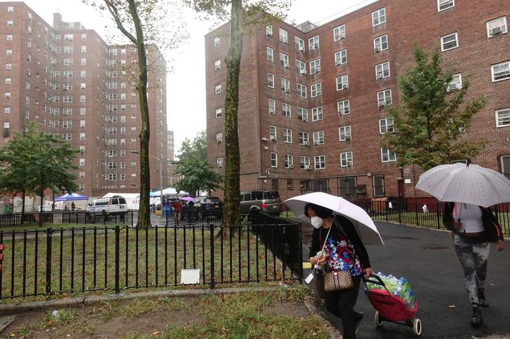 A person holding an umbrella pulls cases of bottled water in a rolling bag outside of the Jacob Riis Houses in the East Village on Tuesday. Another person holding an umbrella is also pictured.